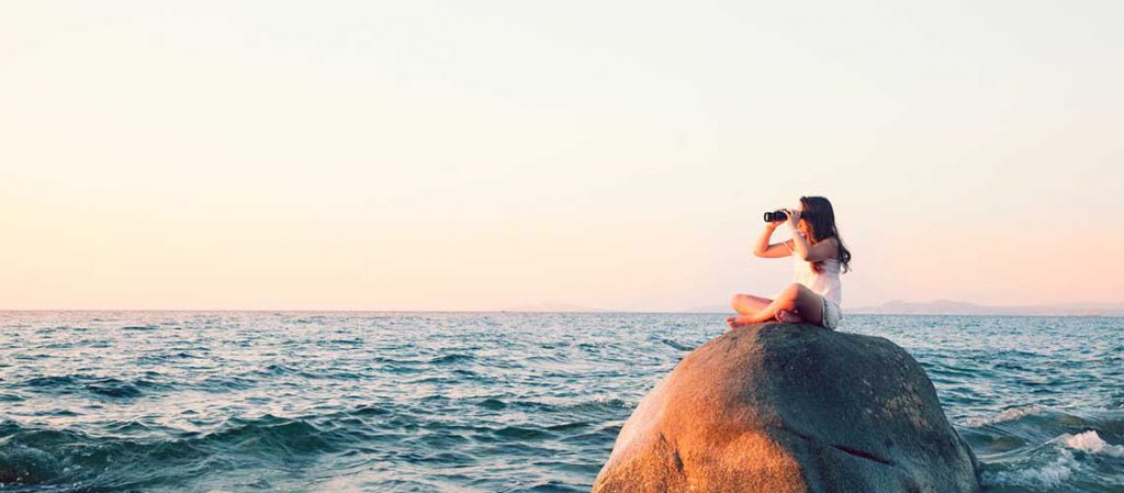 Una joven sentada en una roca frente al mar durante el atardecer, mirando a lo lejos con binoculares. La escena simboliza la planificación y la visión a futuro, representando la importancia del ahorro a largo plazo