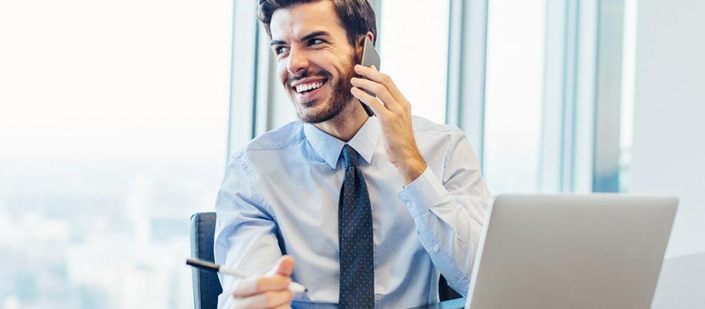 Hombre joven en traje, sonriendo mientras habla por teléfono en una oficina con vista a la ciudad, representando a un asesor financiero ofreciendo orientación y consejos