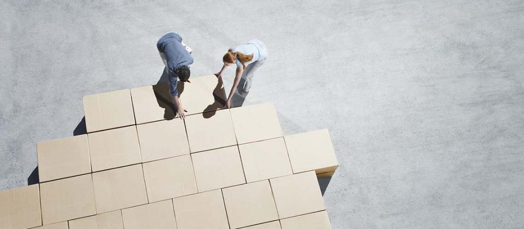 Dos personas colocando grandes cajas de cartón en el suelo, simulando el proceso de construir una casa desde sus cimientos