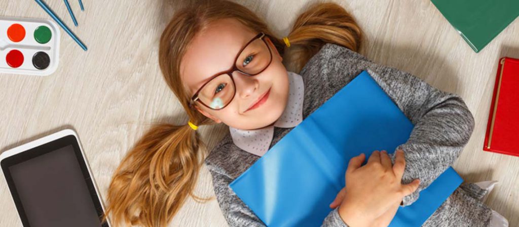 Niña sonriente con gafas, acostada en el suelo con una carpeta azul, rodeada de útiles escolares como tabletas y libros, representando el concepto de ahorro para los estudios de tu hijo