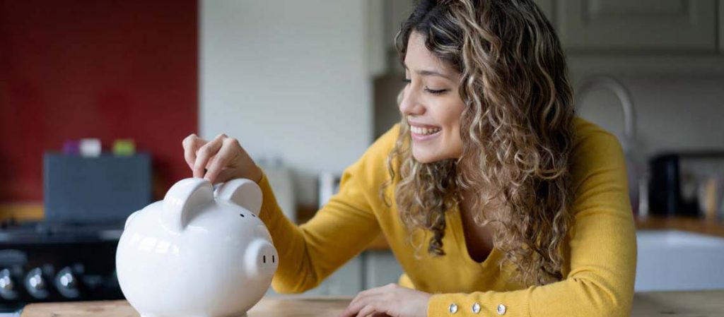 Mujer joven sonriendo mientras deposita una moneda en una hucha de cerdito blanco, representando el concepto de ahorro, relacionado con los SIALP