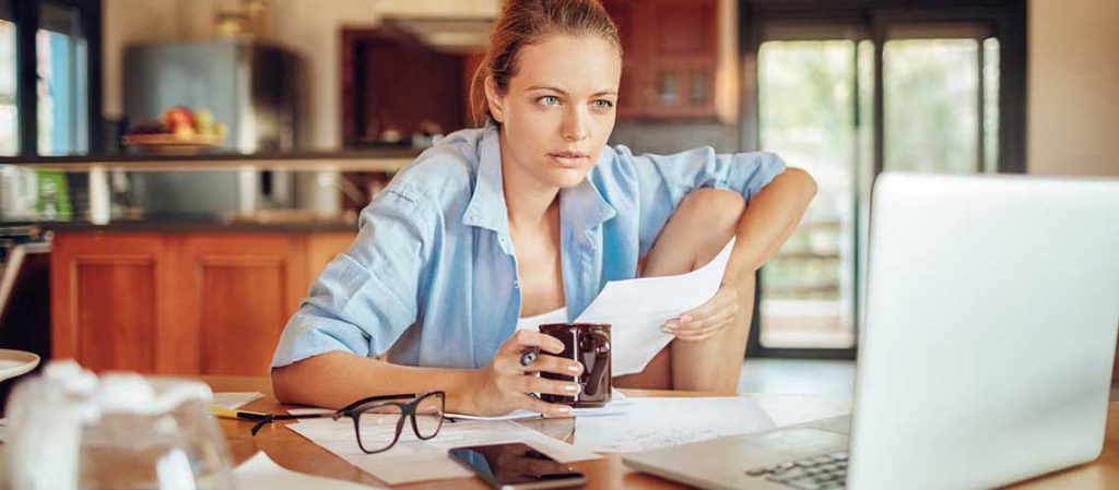 Mujer joven en casa, revisando documentos y tomando notas frente a un ordenador, mientras sostiene una taza. Representa el análisis de los tramos del IRPF